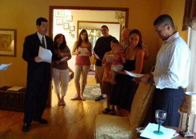 Rabbi Ron Broden presides over a baby naming ceremony in a home, while the father reads along and the mother holds the baby. Family members gather around to celebrate.