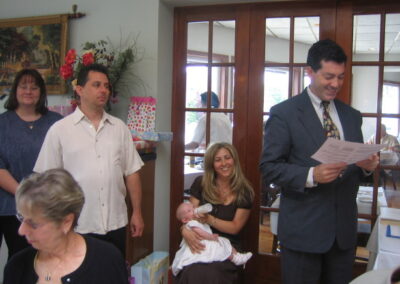 Rabbi Cantor Ron Broden holds a baby naming certificate while standing next to a mother and her newborn baby with family surrounding.