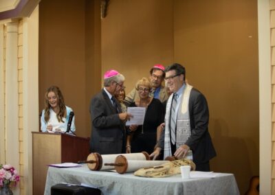 A young girl stands at the bimah, holding a Torah scroll, while Rabbi Cantor Ron Broden leads a Bat Mitzvah ceremony.